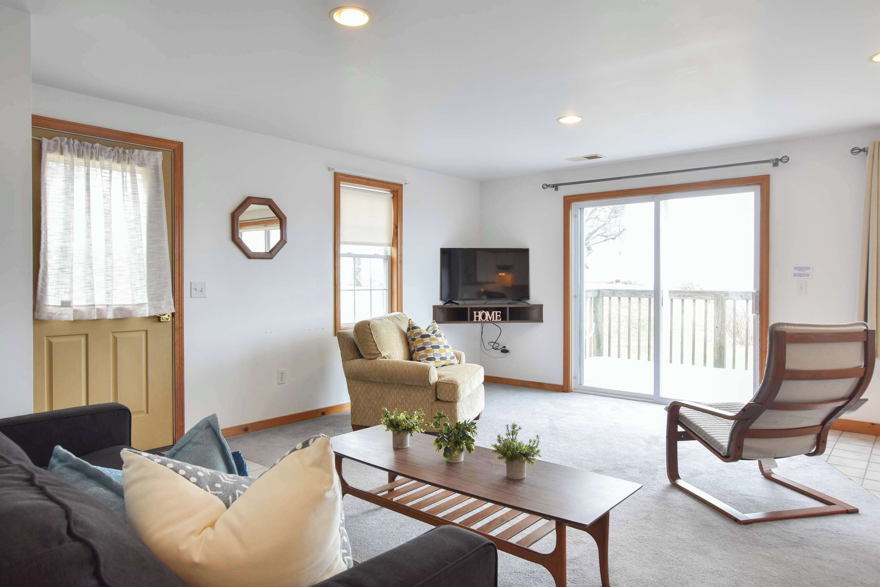 A living room with a sofa, a coffee table with plants, a beige armchair, a wooden chair, a TV mounted on the wall, and a sliding glass door leading to an outdoor area.