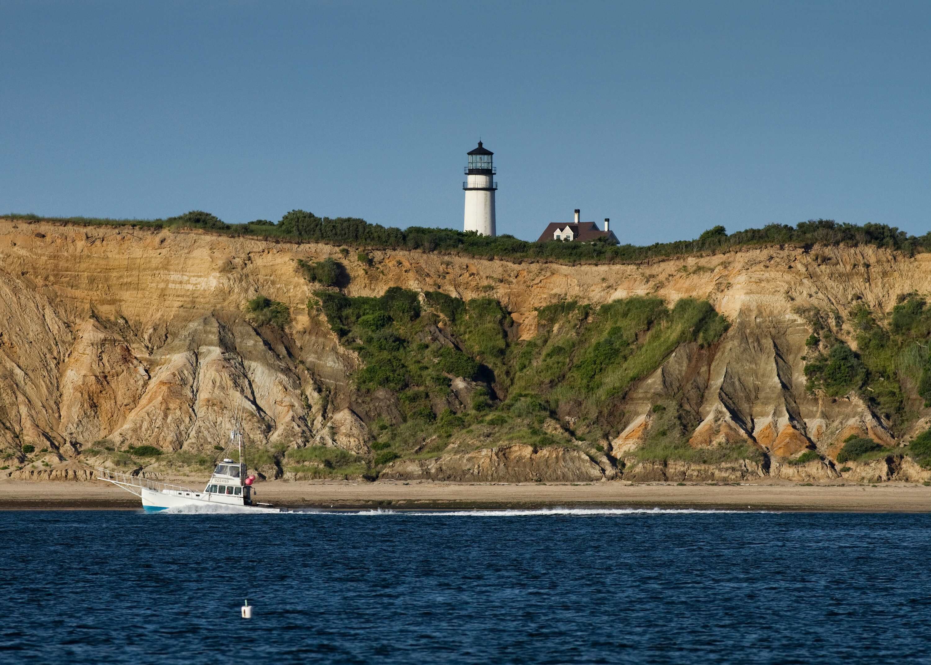 Un paysage côtier avec un phare et une maison perchée sur une falaise, un bateau sur l’eau et un ciel dégagé.