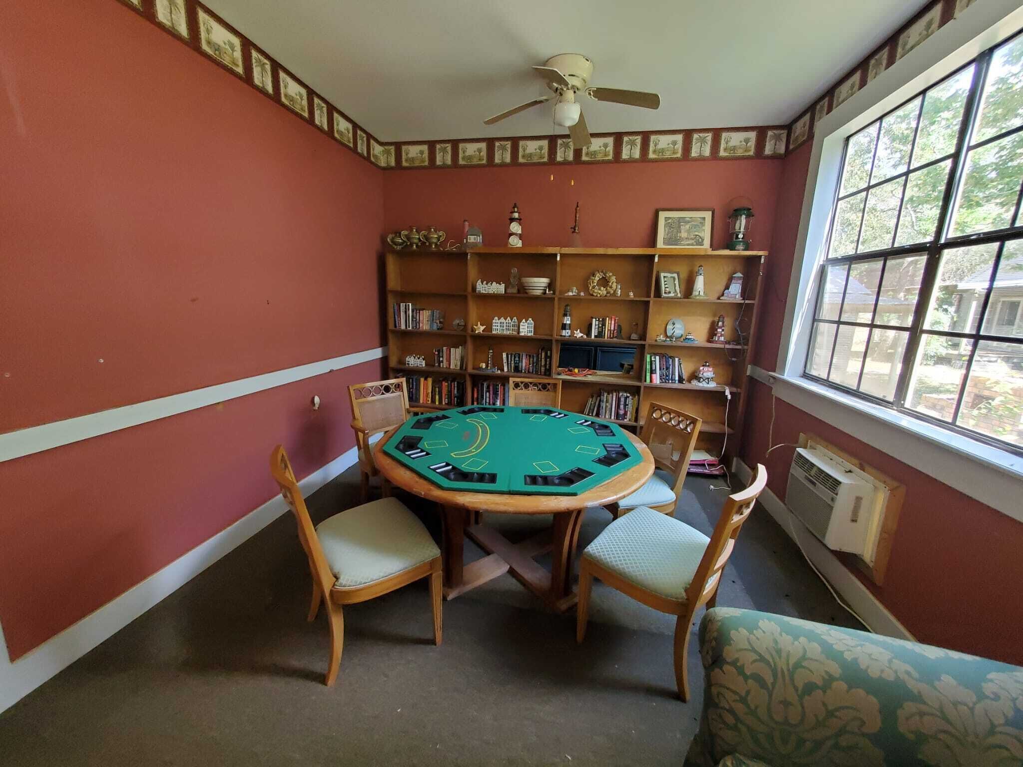 A room with a green felt blackjack table, wooden chairs, a ceiling fan, and a bookshelf filled with books and decorative items.