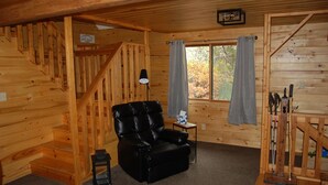 Ein Holzblockhaus-Innenraum mit einem schwarzen Ledersessel, einem kleinen Tisch mit Lampe und einem Fenster mit Vorhängen.