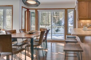 A spacious dining area with a wooden table and chairs, large windows with a view of a snowy deck, and a kitchen with wooden cabinets. - Perfect Family Getaway! (Mont-Tremblant)