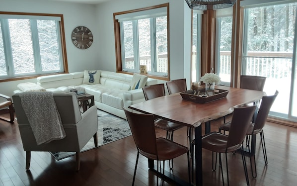 A spacious living room with a white sofa, a wooden dining table with chairs, and large windows offering a view of snow-covered trees. - Perfect Family Getaway! (Mont-Tremblant)