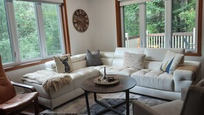 Ein Wohnzimmer mit einem weißen Sofa, einem braunen Ledersessel, einem Couchtisch und einem großen Fenster mit Blick auf Bäume.