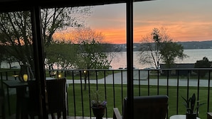 A balcony with a view of a sunset over a body of water, surrounded by trees and a grassy area.