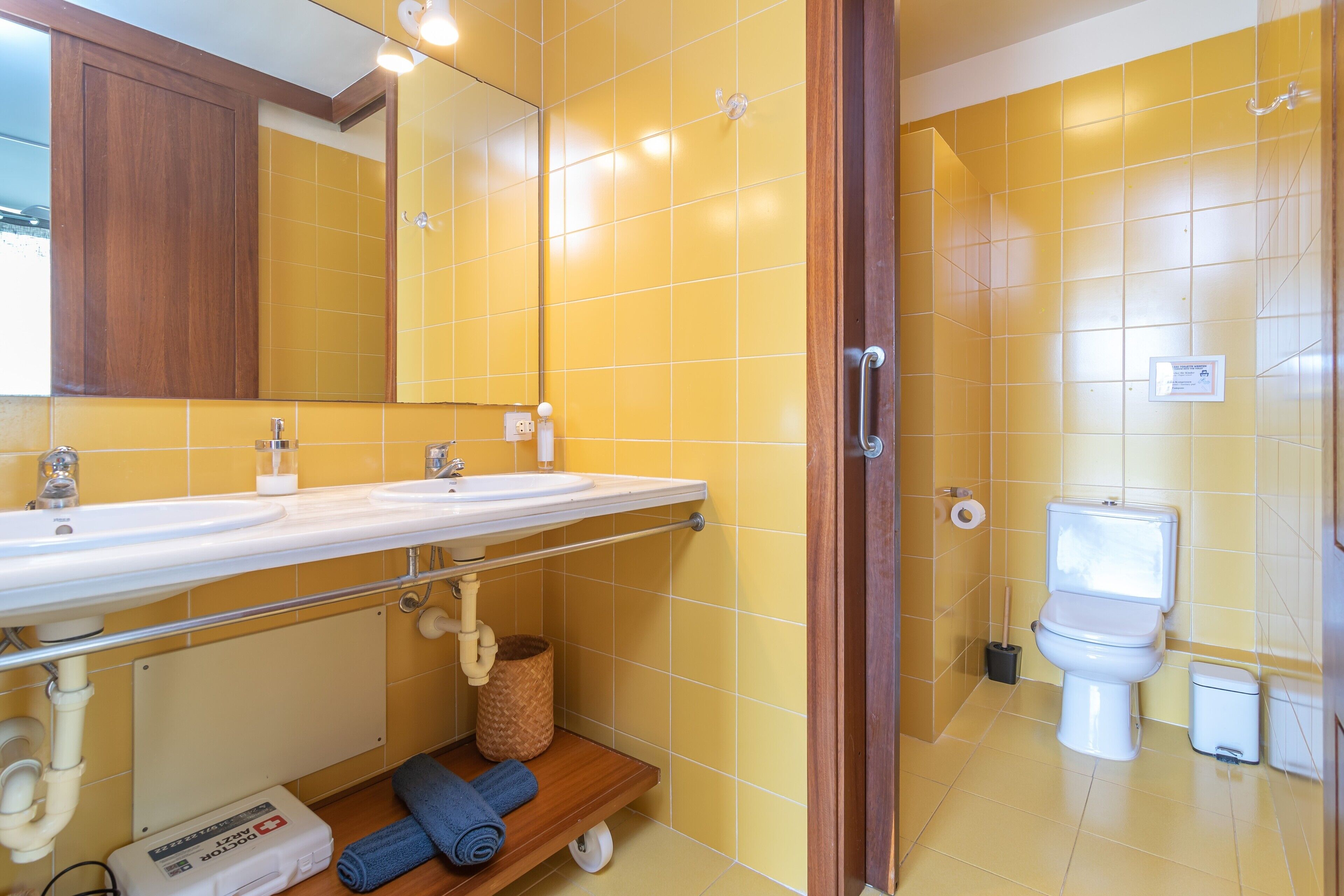 A bathroom with yellow tiles, a white sink, a mirror, a toilet, and a blue towel on a shelf.