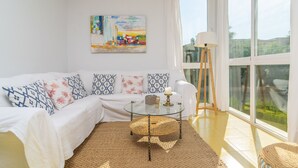 A modern living room with a white sofa, patterned cushions, a glass coffee table, and a large window with curtains.