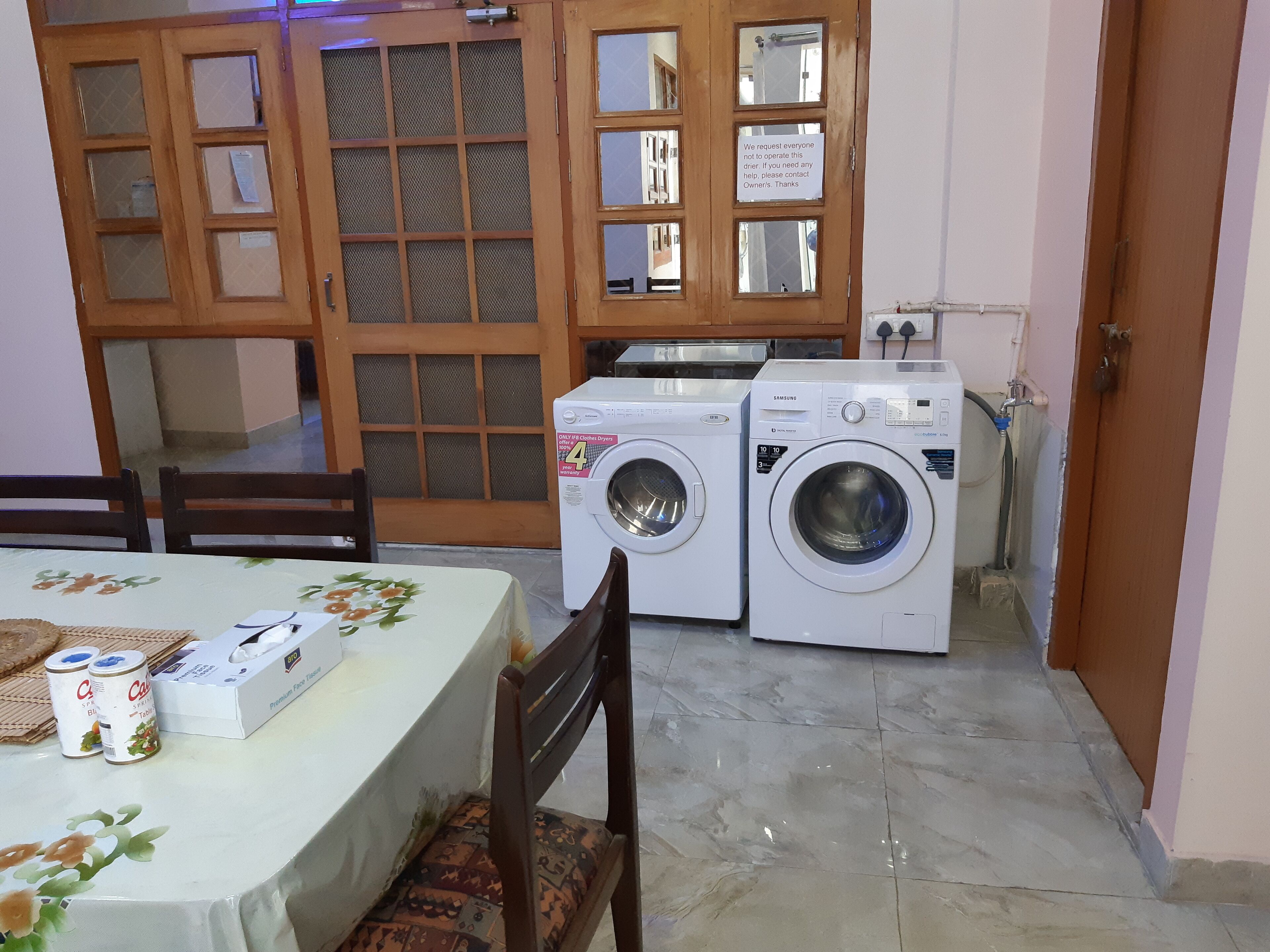 A laundry area with two washing machines, a table with a floral tablecloth, and a wooden door with a sign.