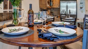 Mesa de comedor dispuesta para cuatro, con copas de vino, platos y cubiertos. Una botella de vino está sobre la mesa, y hay flores en un jarrón. En el fondo, se observan armarios de madera y un refrigerador.