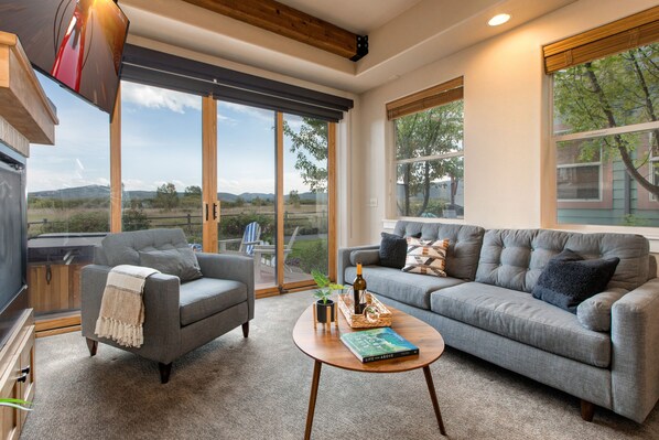 A modern living room with a grey sofa, a grey armchair, and a coffee table. There is a large window with a view of the outdoors.