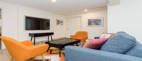 A modern living room with a flat-screen TV, orange and blue sofas, a coffee table, and artwork on the walls.