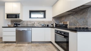 A modern kitchen with a dishwasher, oven, microwave, and a window above the sink. - Across The Road From The Beach (Inverloch)