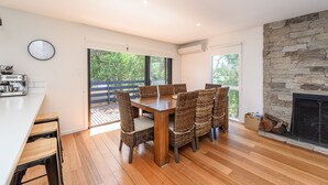 Un espace repas moderne avec une table en bois et des chaises en osier, de grandes fenêtres donnant sur un paysage verdoyant et un mur en pierre agrémenté d’une cheminée.