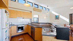 A modern kitchen with wooden cabinets, a built-in oven, a dishwasher, and a large island with a sink. There are multiple windows providing natural light and a view of the outside. - Picture Perfect on Pier (INVERLOCH)