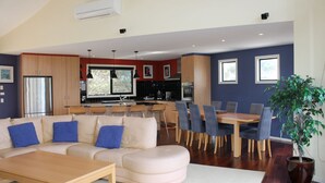 A modern living room with a sofa, coffee table, and dining area with chairs. There is a kitchen with a refrigerator, stove, and sink. A large window provides natural light. - Stunning Sea Views on Surf (CAPE PATERSON)