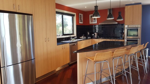 A modern kitchen with wooden cabinets, stainless steel appliances, a black countertop, and bar stools. - Stunning Sea Views on Surf (CAPE PATERSON)