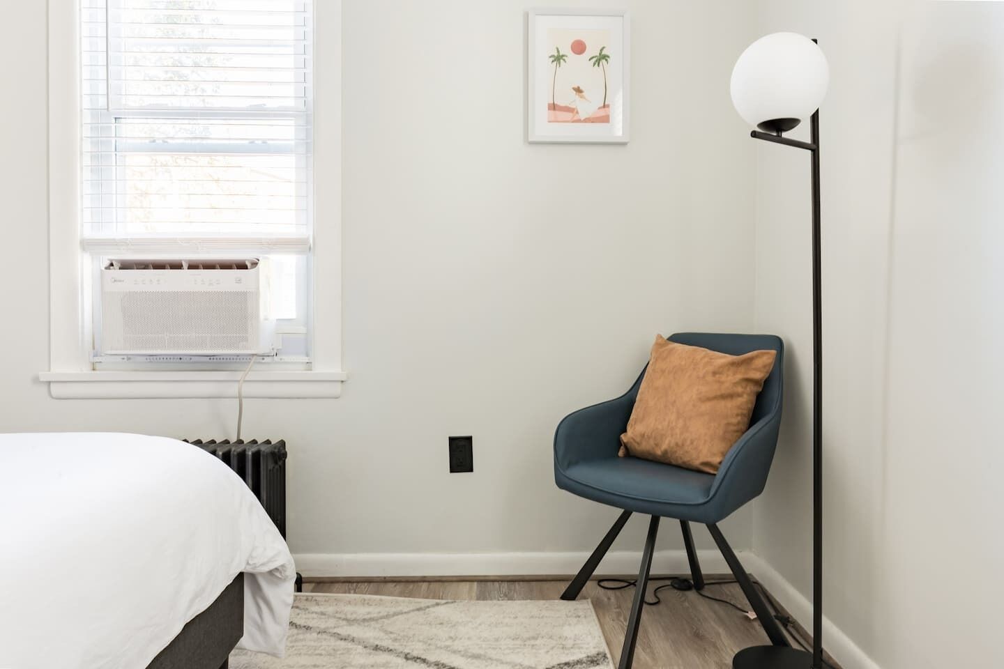 A bedroom with a bed, a chair with a cushion, a floor lamp, a radiator, and a window with blinds.