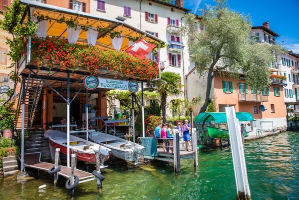 A riverside restaurant with outdoor seating, boats docked nearby, and a clear view of the water. - Sunlight - 150m from the lake, Lugano, Switzerland (Lugano)