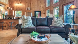 A cozy cabin interior with a wooden interior, a leather sofa, a coffee table with a tray, and a kitchen area with wooden cabinets and a window. - Cozy cabin w/ Hot tub, Arcade, Wi-Fi, Air Hockey & Views (Sevierville)
