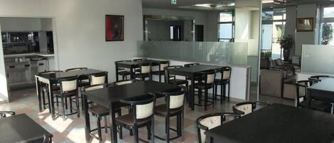 A modern dining area with multiple tables and chairs, a kitchen in the background, and a large window providing natural light.