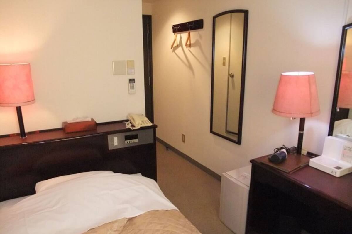 A hotel room with a bed, bedside lamps, a telephone, a mirror, a small desk with a lamp, and a trash bin.