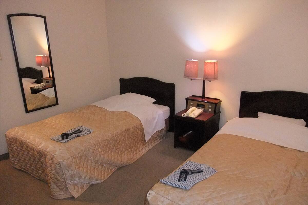 A hotel room with two single beds, a nightstand with a phone, a lamp, and a mirror on the wall.