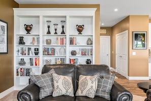 A living room with a leather sofa, a white bookshelf filled with books and decorative items, and a framed picture on the wall. - 🛶 Golf in Luxury | Torch Lake Getaway at A-Ga-Ming | 2 En-Suites ➠ 12534 (Kewadin)