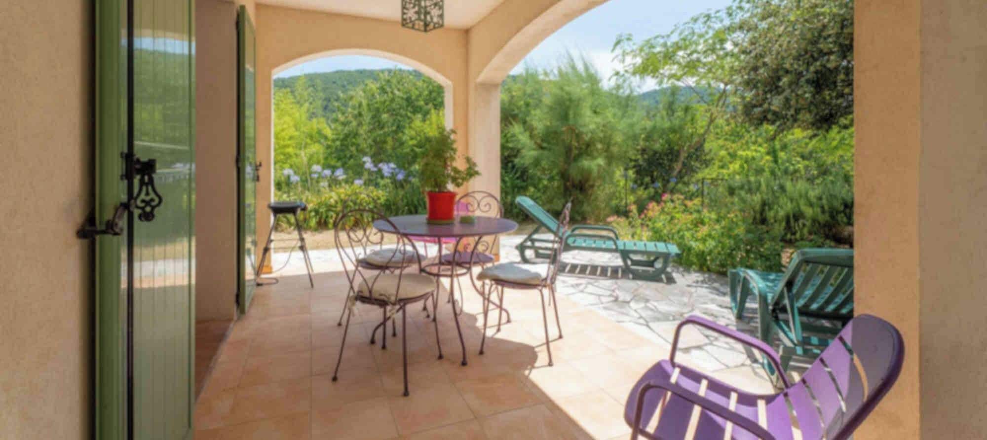 Uma área de varanda coberta com mesa e cadeiras, uma porta verde e vista para um jardim exuberante.