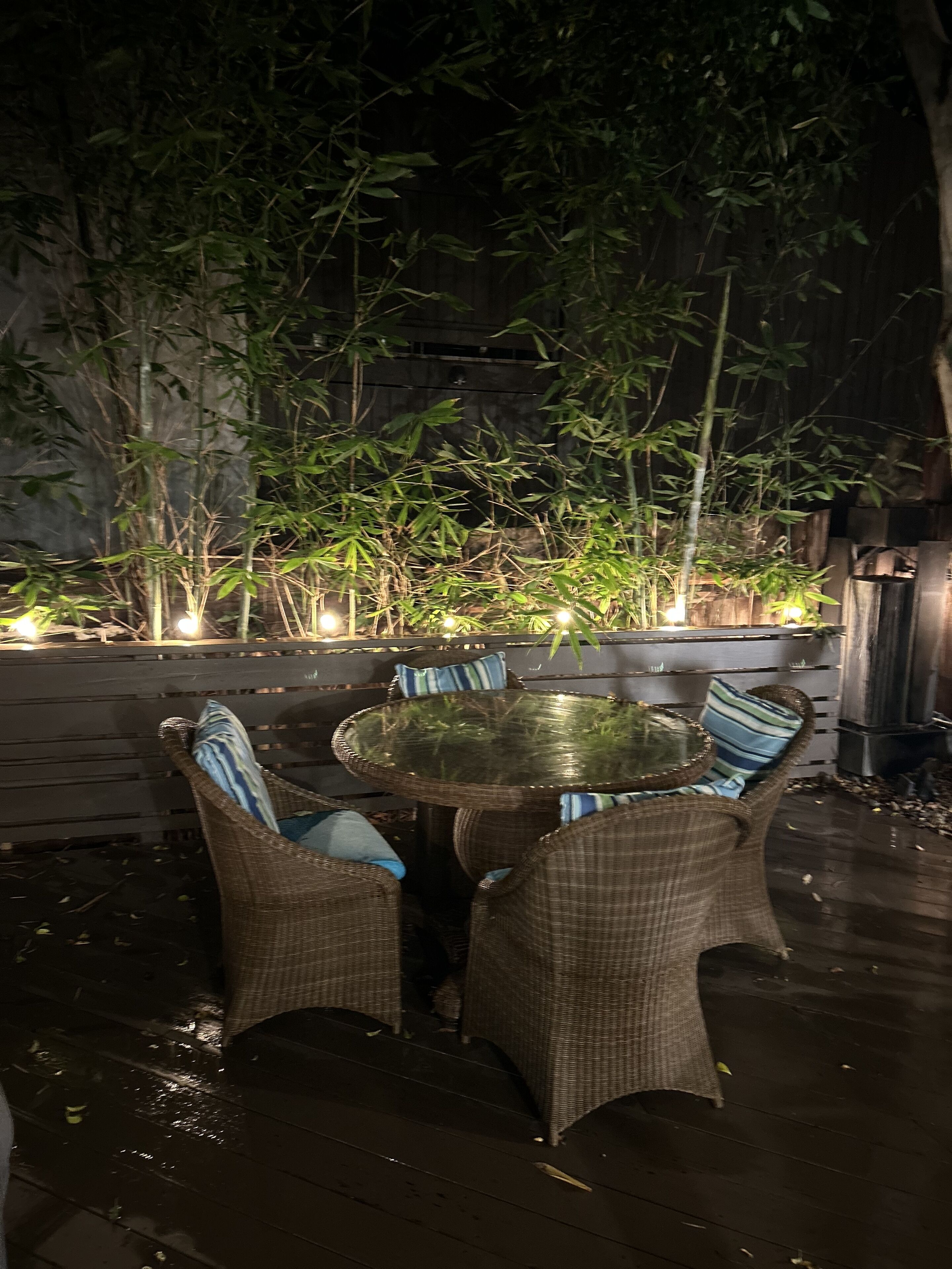 An outdoor dining area with wicker furniture and a glass table, surrounded by bamboo plants and a wooden fence.