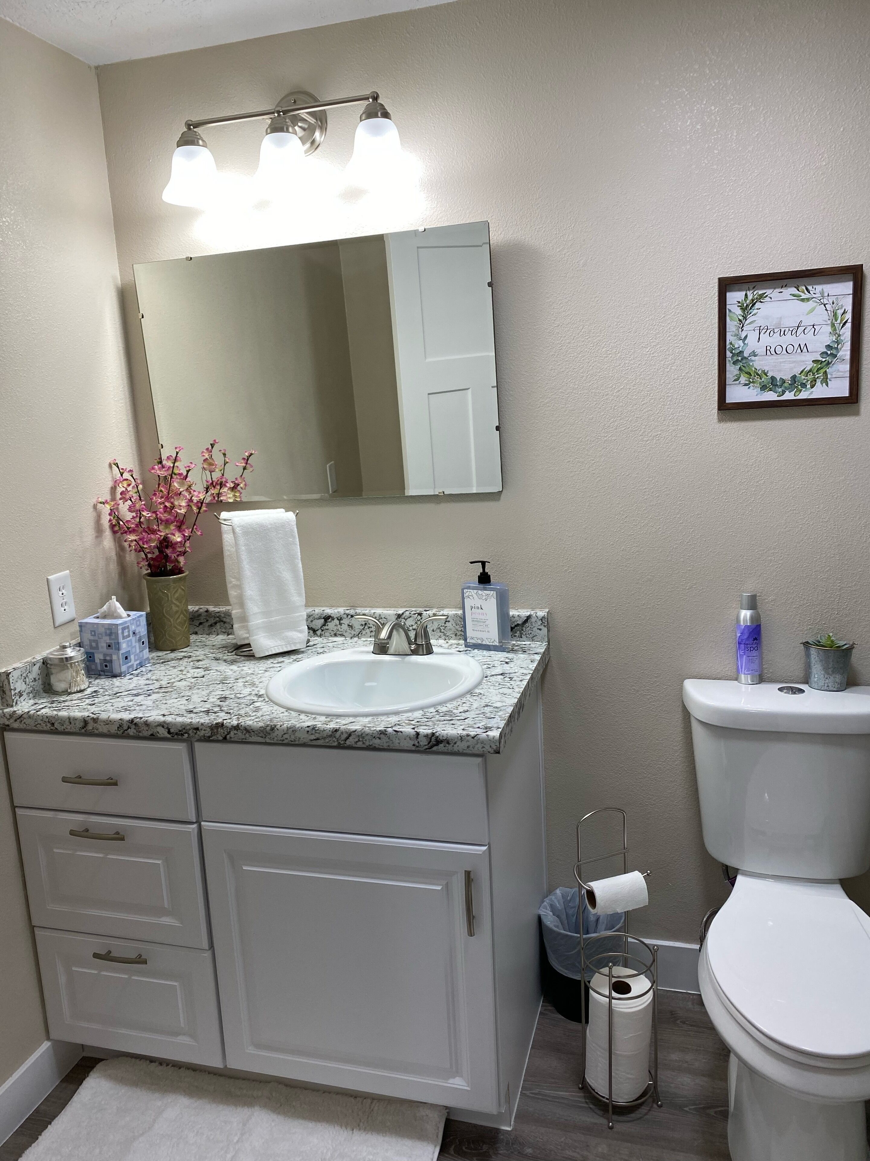 A bathroom with a white sink, vanity, and mirror. There is a framed sign on the wall, a toilet, and a trash can.