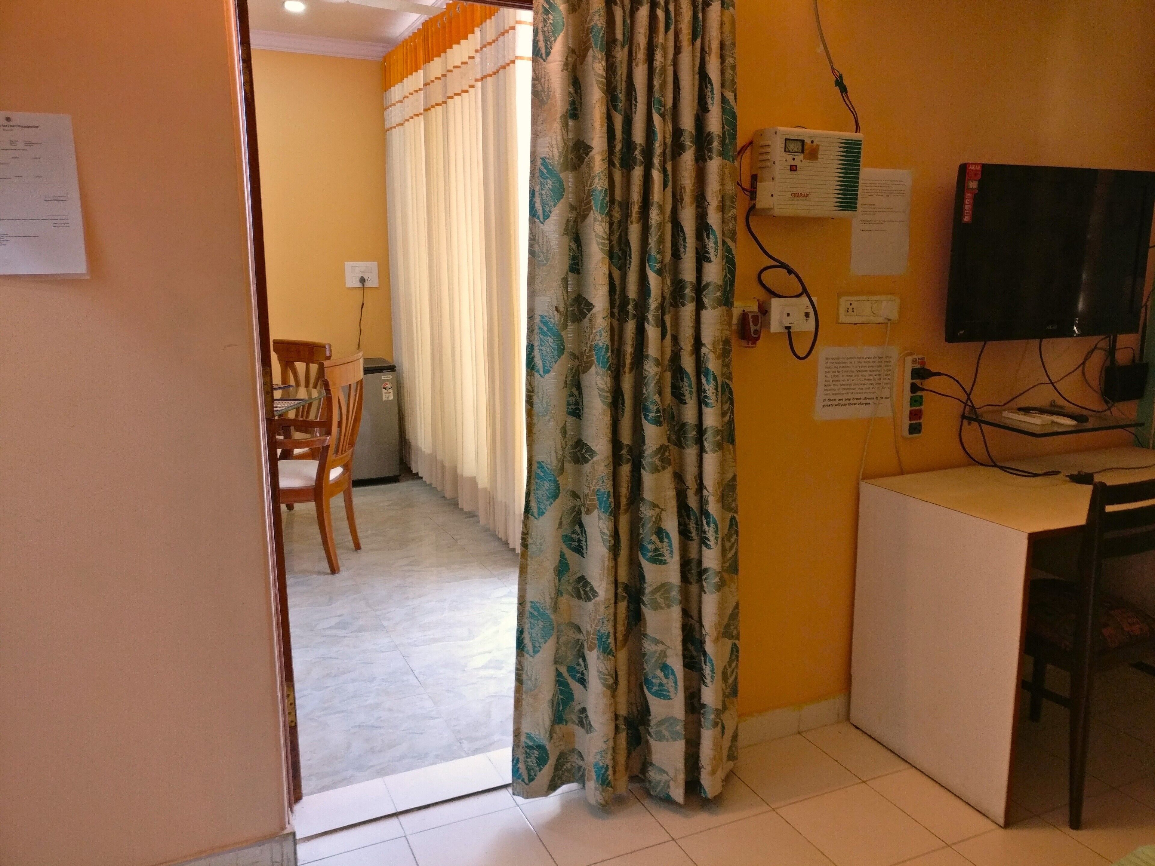 Una stanza con una scrivania, una sedia e una televisione. Si vede una tenda con un motivo a foglia e una presa elettrica sulla parete.