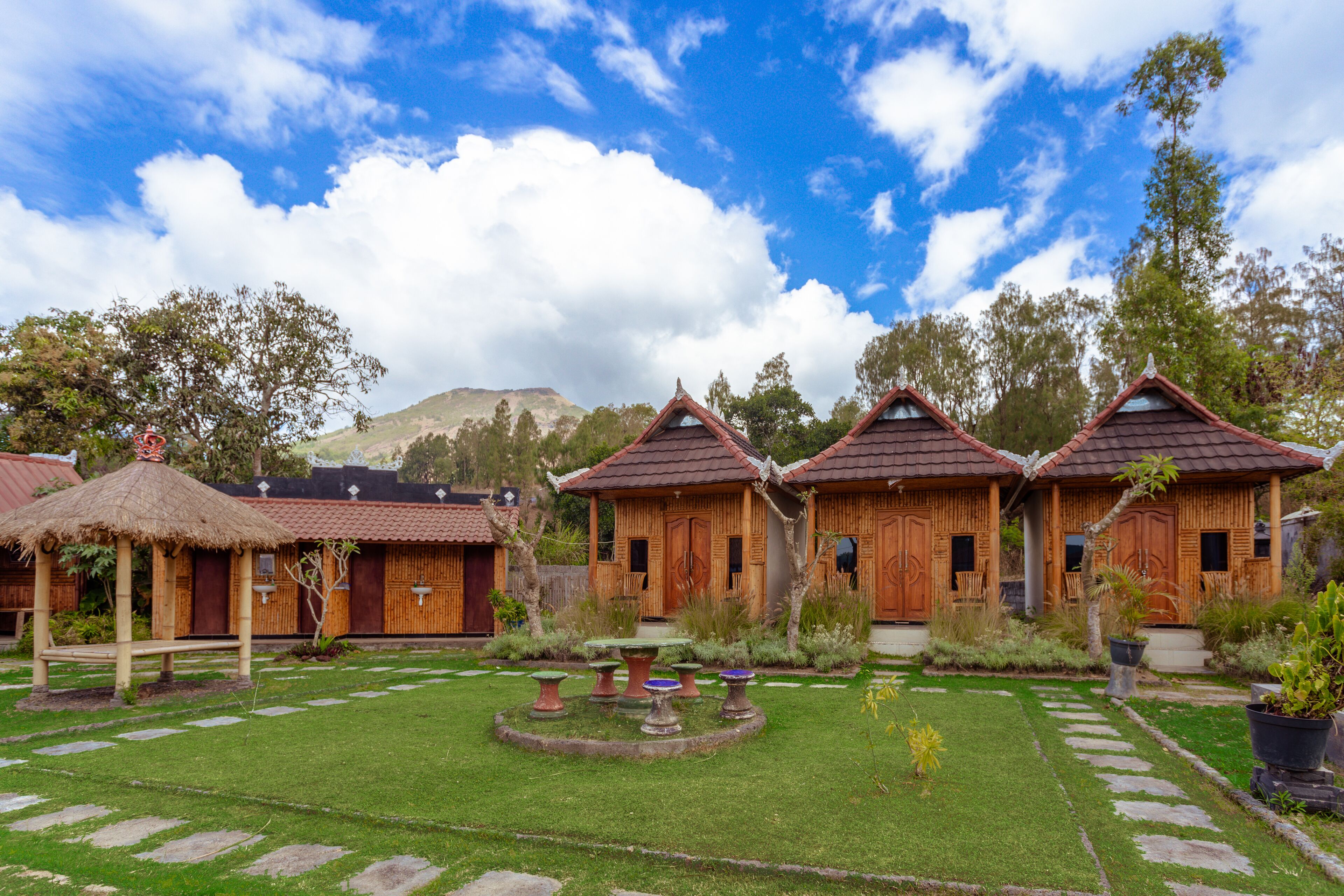 Un patio con casas de madera, una construcción con techo de paja y una montaña al fondo.
