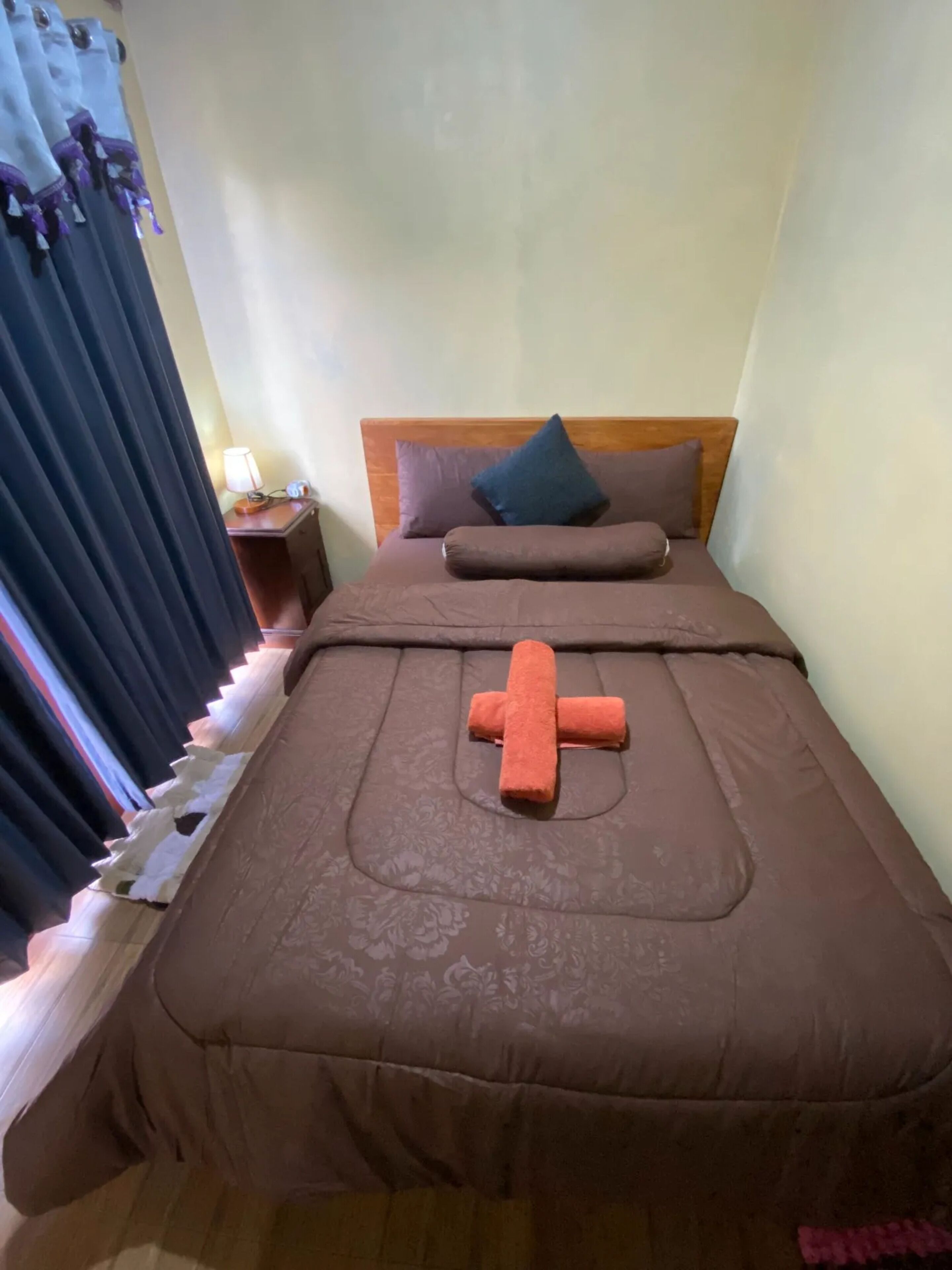 Una cama bien tendida con una colcha marrón y una almohada con forma de cruz.