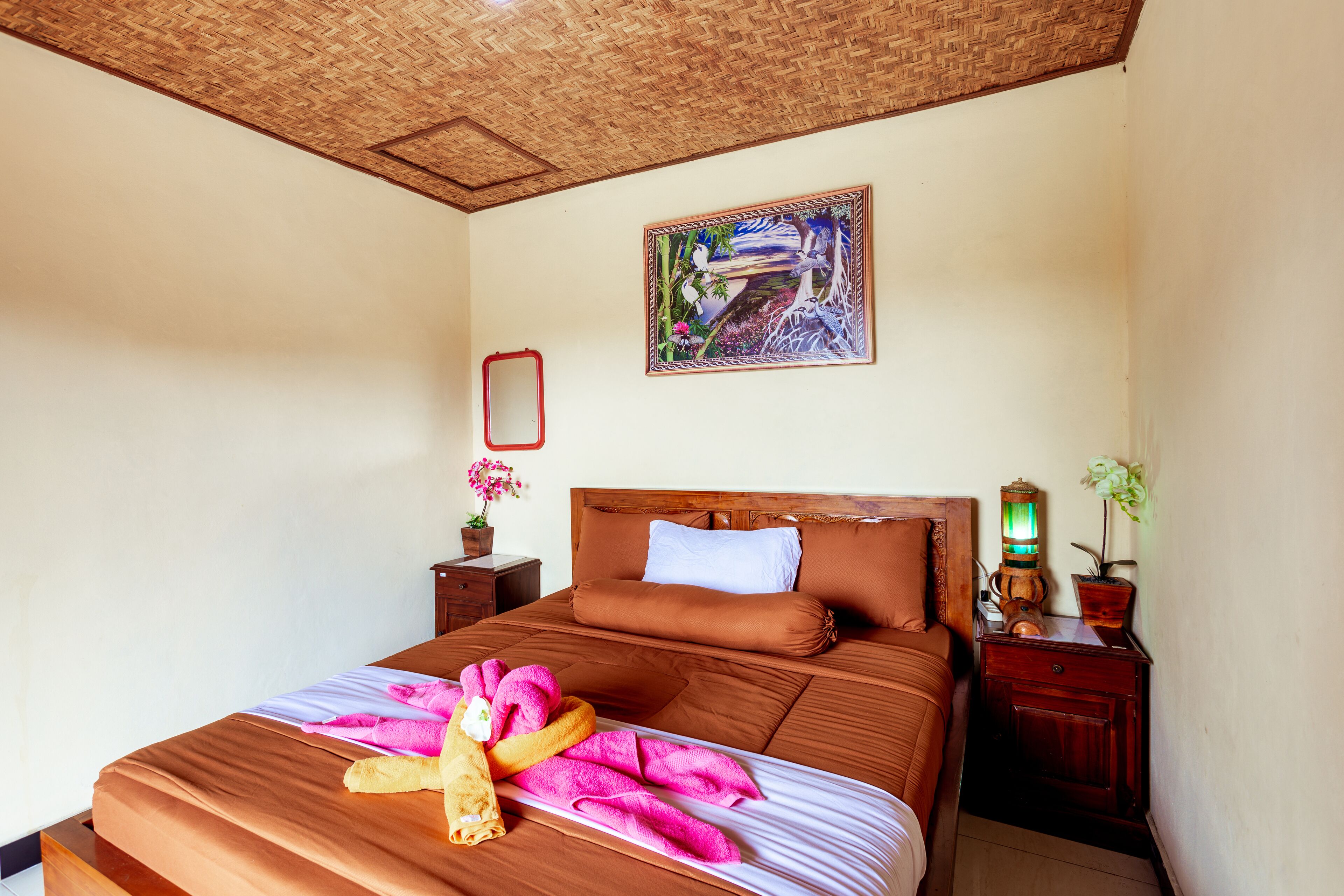 Un lit bien fait, agrémenté de serviettes roses et d’une serviette jaune, une tête de lit en bois, une table de chevet avec une lampe et un tableau encadré au mur.