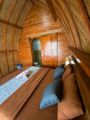 A cozy, wooden room with a bed, pillows, and a small table. - Batur Bamboo Cabin (Kintamani)