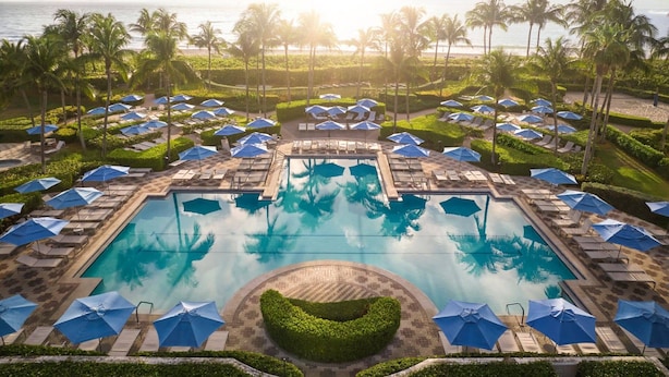 Zwembadgedeelte van een resort met meerdere ligbedden en parasols, omringd door palmbomen en uitzicht op de oceaan.