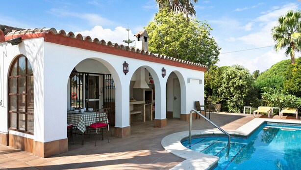 Un espace aménagé au bord de la piscine, avec un bâtiment blanc doté d’arcades, une terrasse carrelée et une piscine.