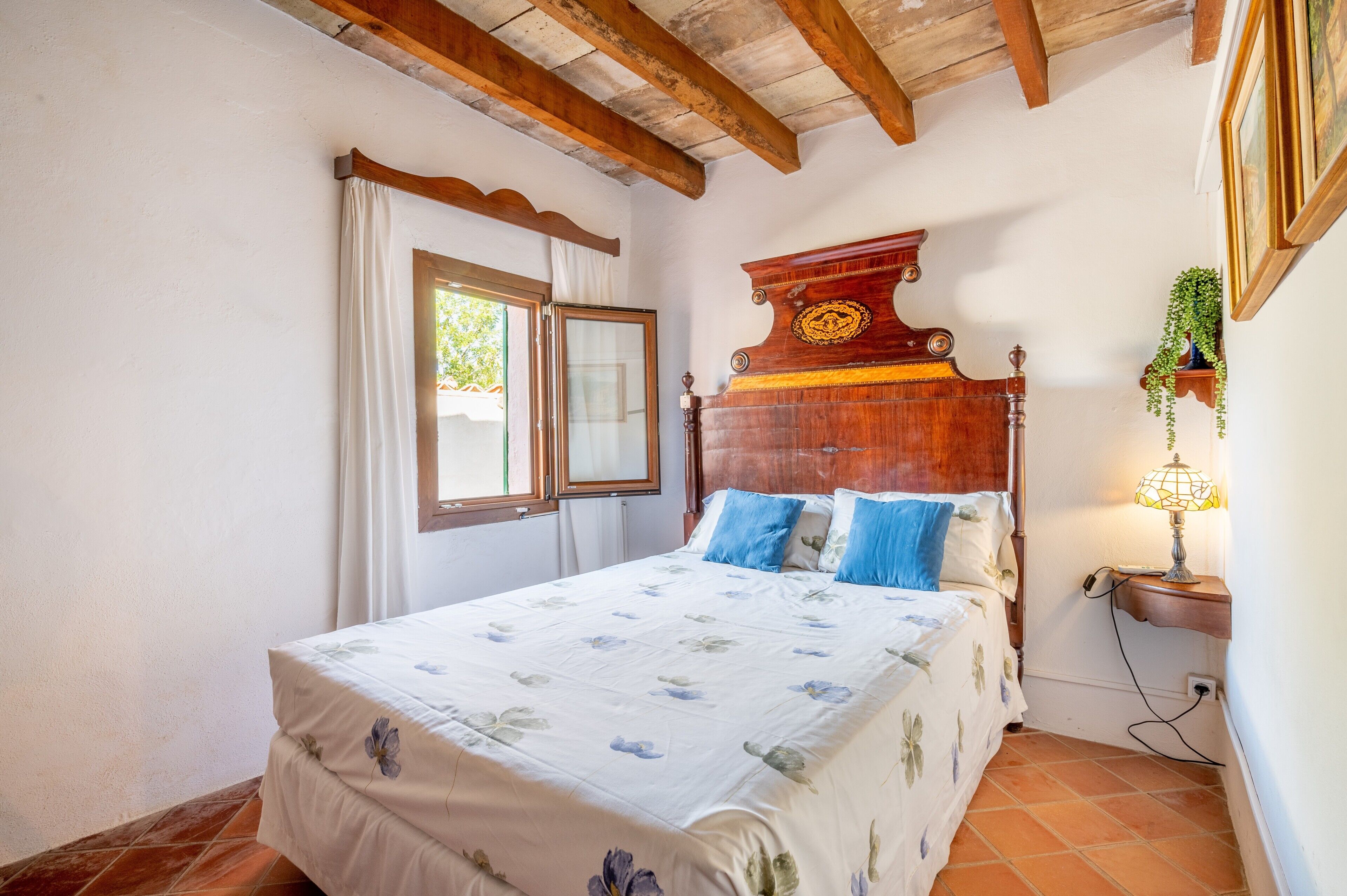 Un dormitorio con estructura de cama de madera, una ventana con cortinas, una lámpara de noche y un estante en la pared con una planta.