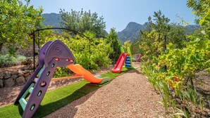 A playground with slides and swings, surrounded by greenery and a mountain backdrop.