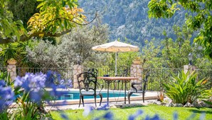 A poolside area with a table and chairs, surrounded by greenery and a mountain backdrop.