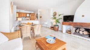 A living room with a fireplace, wicker furniture, and a kitchen in the background. - Holiday Apartment Montemar 36 with Sea and Mountain View, Wi-Fi, Pool & Terrace (Port de Soller)