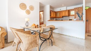 A dining area with wicker chairs, a glass table, and a kitchen with wooden cabinets. - Holiday Apartment Montemar 36 with Sea and Mountain View, Wi-Fi, Pool & Terrace (Port de Soller)