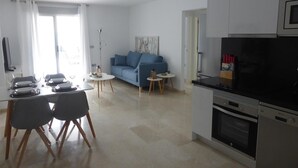Eine moderne Wohnung mit Essbereich, einem Wohnzimmer mit blauem Sofa, einer Küche mit Mikrowelle und Backofen sowie einem großen Fenster.