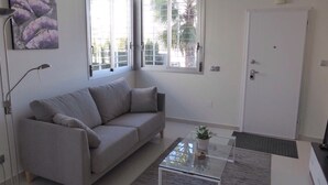 Ein Wohnzimmer mit grauem Sofa, einem Glastisch, einer Tür und Fenstern mit Jalousien.