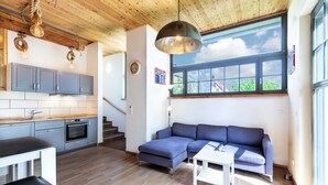 Een modern interieur met keuken, eethoek en zithoek, met houten balkenplafond, een groot raam en een blauwe bank.