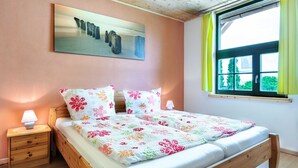 Een tweepersoonsbed met bloemenbeddengoed, een nachtkastje met lamp, een schilderij aan de muur, en een raam met gordijnen.