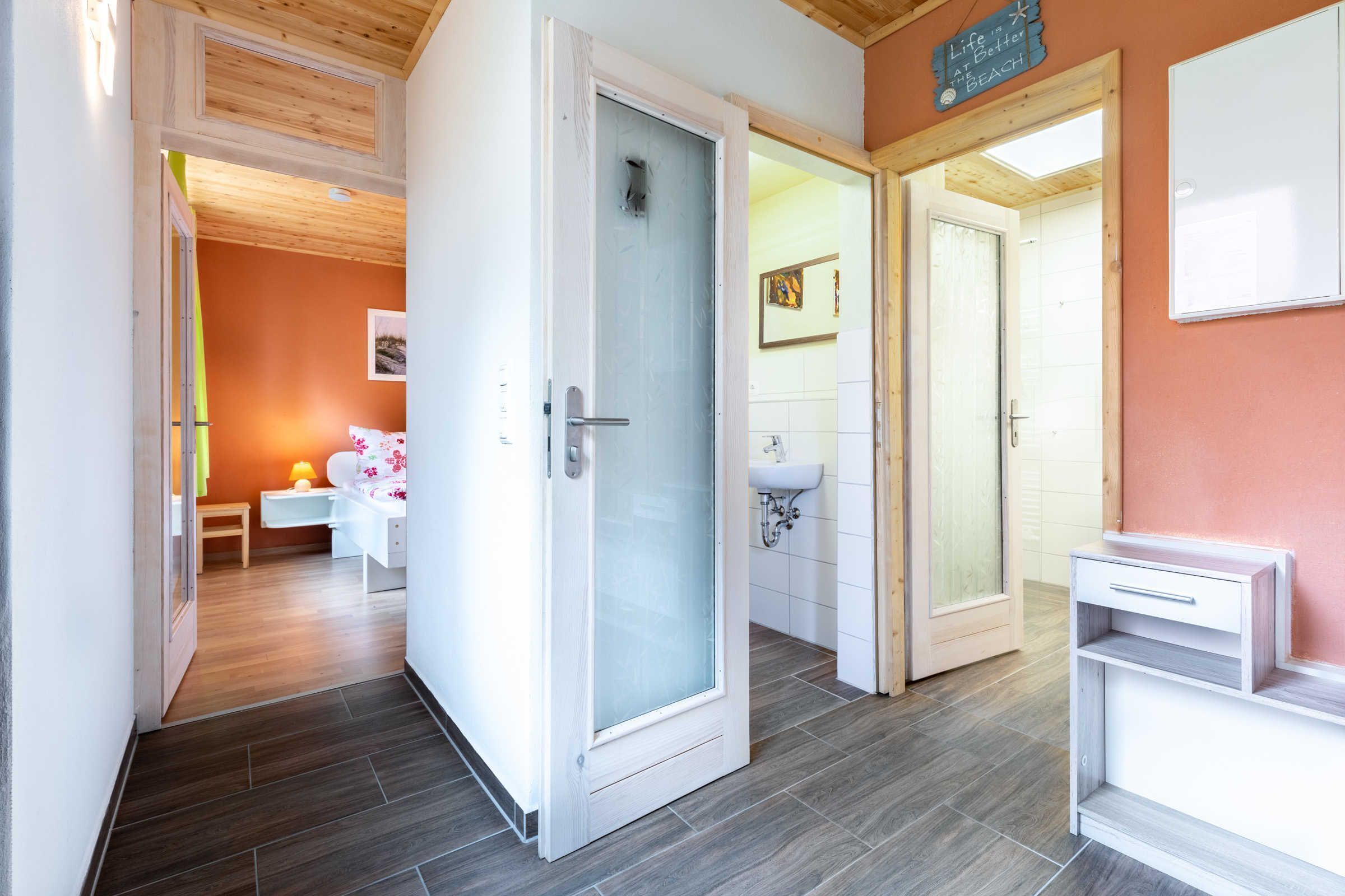 Ein Hotelzimmer mit Holzboden, Glastür, Waschbecken in Weiß und einem Nachttisch mit Lampe.