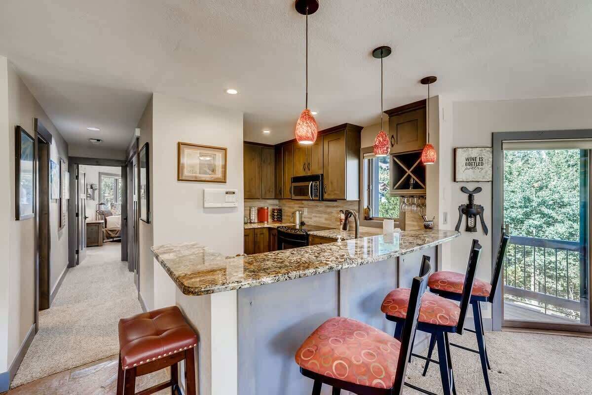 A modern kitchen with a granite countertop, bar stools, pendant lights, and a view of the outdoors.