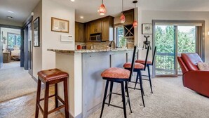 A modern kitchen with a bar area, wooden cabinets, and pendant lighting. There are three bar stools and a red sofa in the adjacent living area. - Platinum Frost Fire, Hot Tub access, Near Slopes! (Keystone)