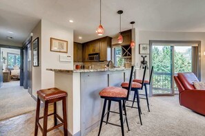 A modern kitchen with a bar area, wooden cabinets, and pendant lighting. There are three bar stools and a red sofa in the adjacent living area.