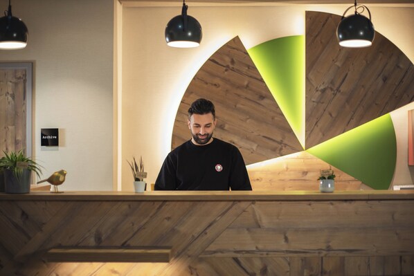 A reception desk with a wooden finish, a black and white sign, and a green accent wall. - Holiday Inn – the niu, Mill Cologne Mülheim by IHG (Cologne)